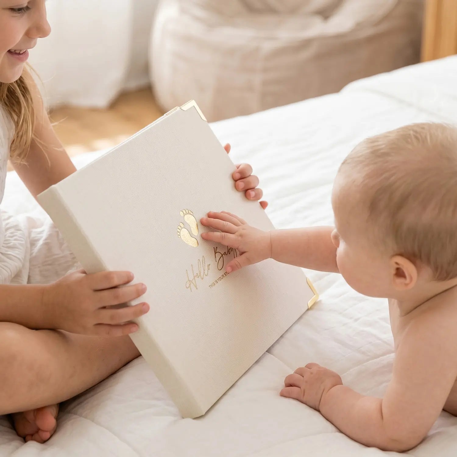 A baby reaching out to touch the Hello Baby memory book held by an older sibling, highlighting the premium gender-neutral linen cover, delicate gold foil footprint stamping, and elegant metal corners of this family heirloom.