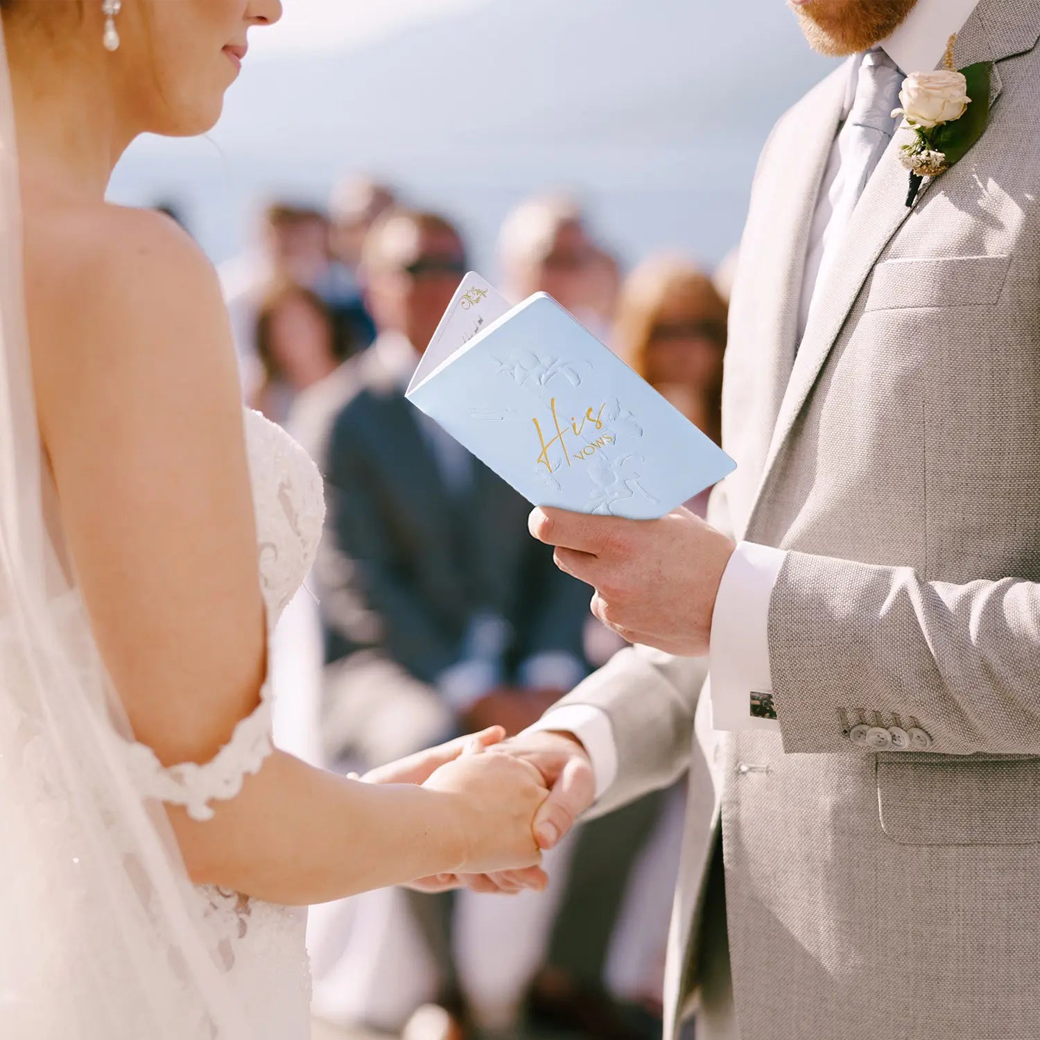 A groom in a light grey suit reading from the Molekaus light blue embossed 'His Vows' book at the altar, holding the bride's hand during their wedding ceremony vow exchange.